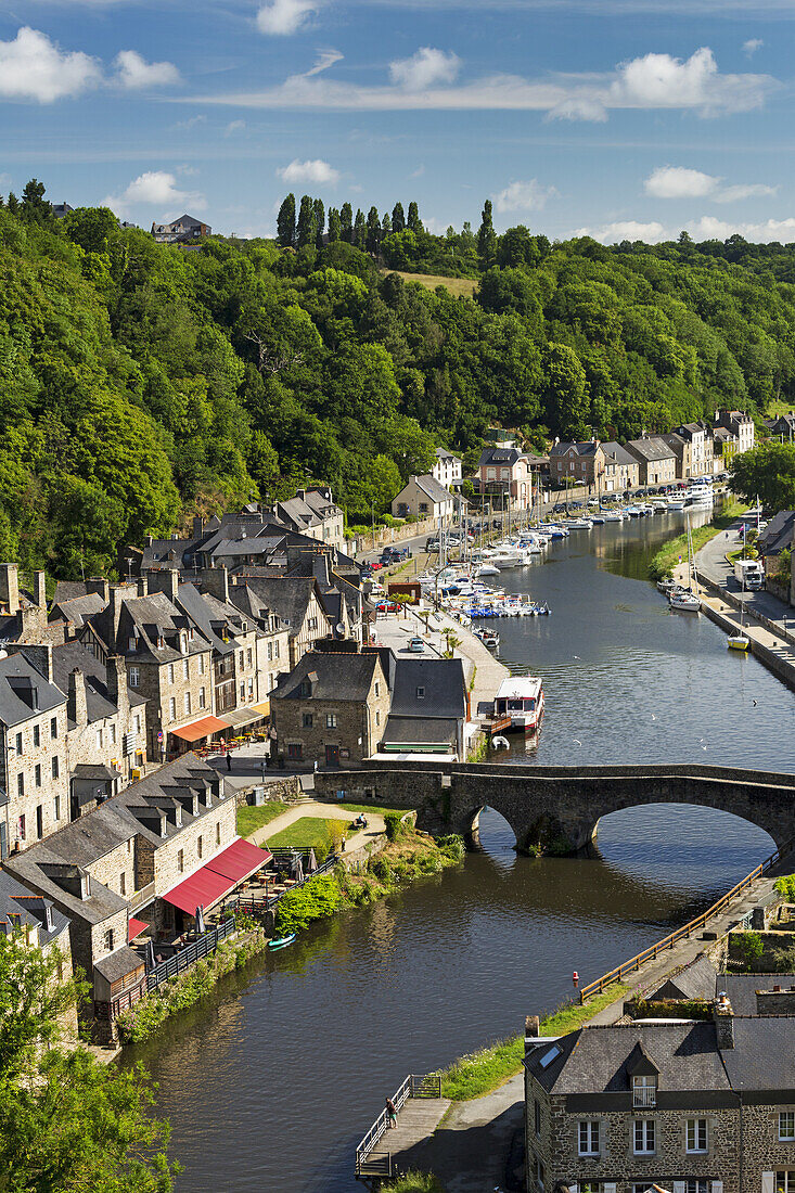 Baumbewachsene Talstadt am Flussufer, Steinbrücke und Boote im Hafen mit blauem Himmel und Wolken; Dinan, Bretagne, Frankreich