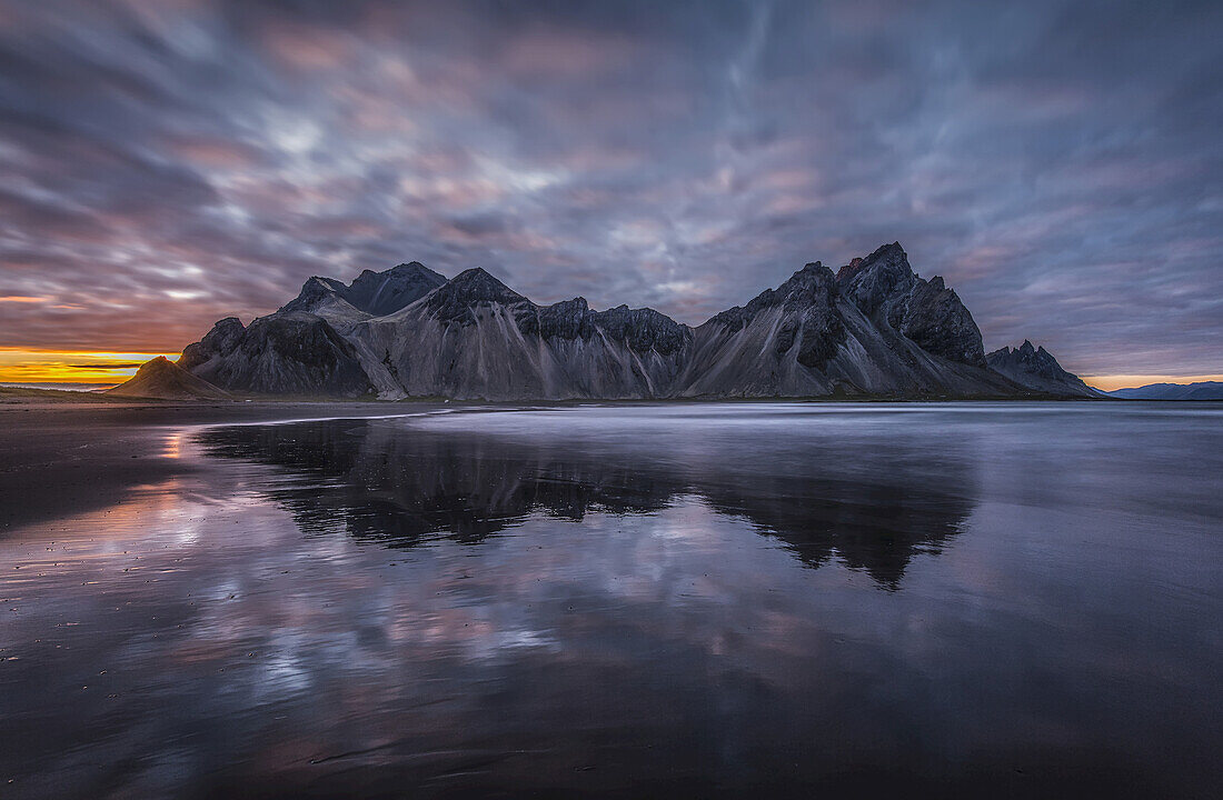 Rugged Mountain Peaks And A Colourful Sunset Reflected In Tranquil Water; Iceland