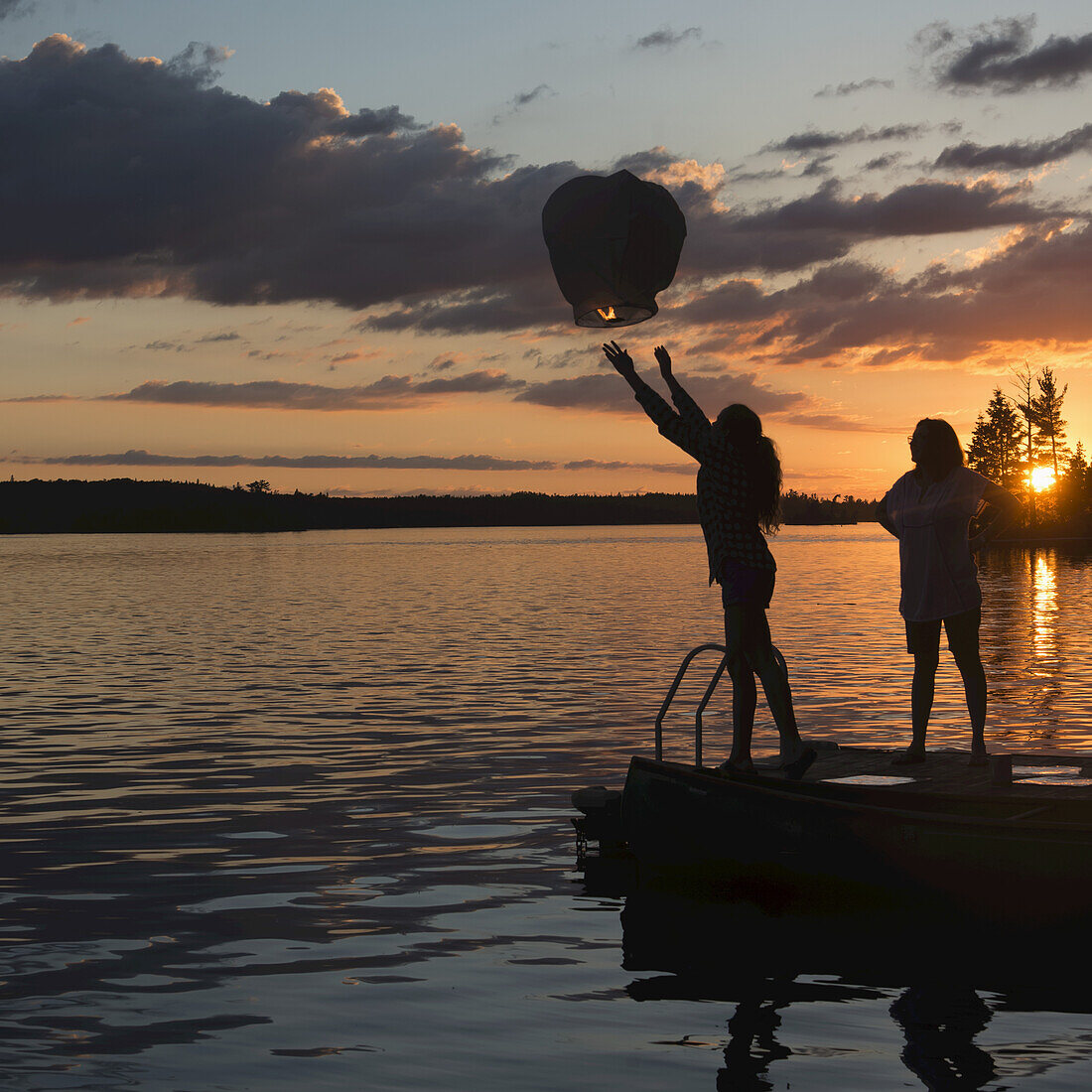 Silhouette Of A Teenage Girl Releasing A Lit Lantern Out Over A Lake At Sunset; Lake Of The Woods, Ontario, Canada