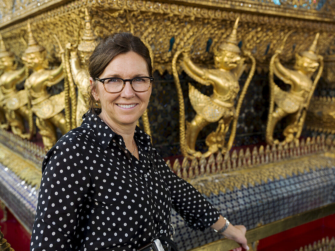 Porträt einer Frau, die neben einer Wand mit Goldstatuen steht, Tempel des Smaragdbuddhas (Wat Phra Kaew); Bangkok, Thailand