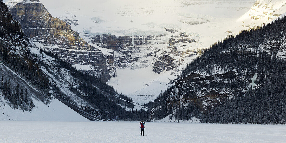 A Person Standing On Frozen Lake Louise Taking A Picture Of The Snow Covered Mountains; Lake Louise, Alberta, Canada