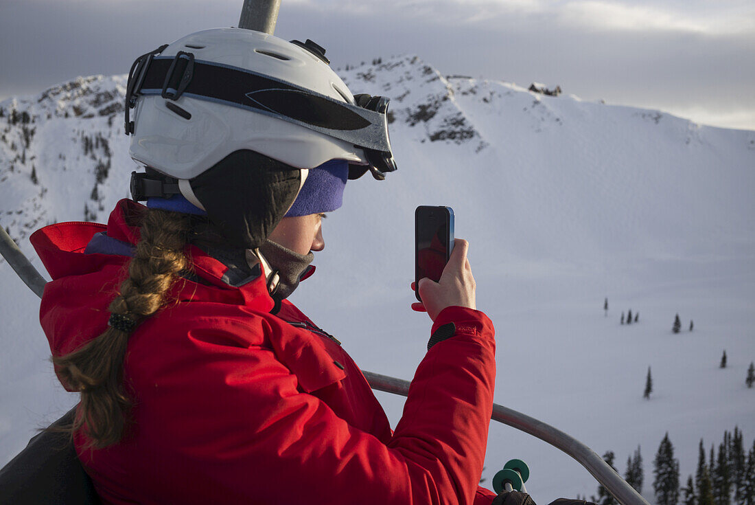 Ein Skifahrer auf einem Sessellift in einem Skigebiet macht ein Foto mit einem Smart Phone, Banff National Park; Alberta, Kanada