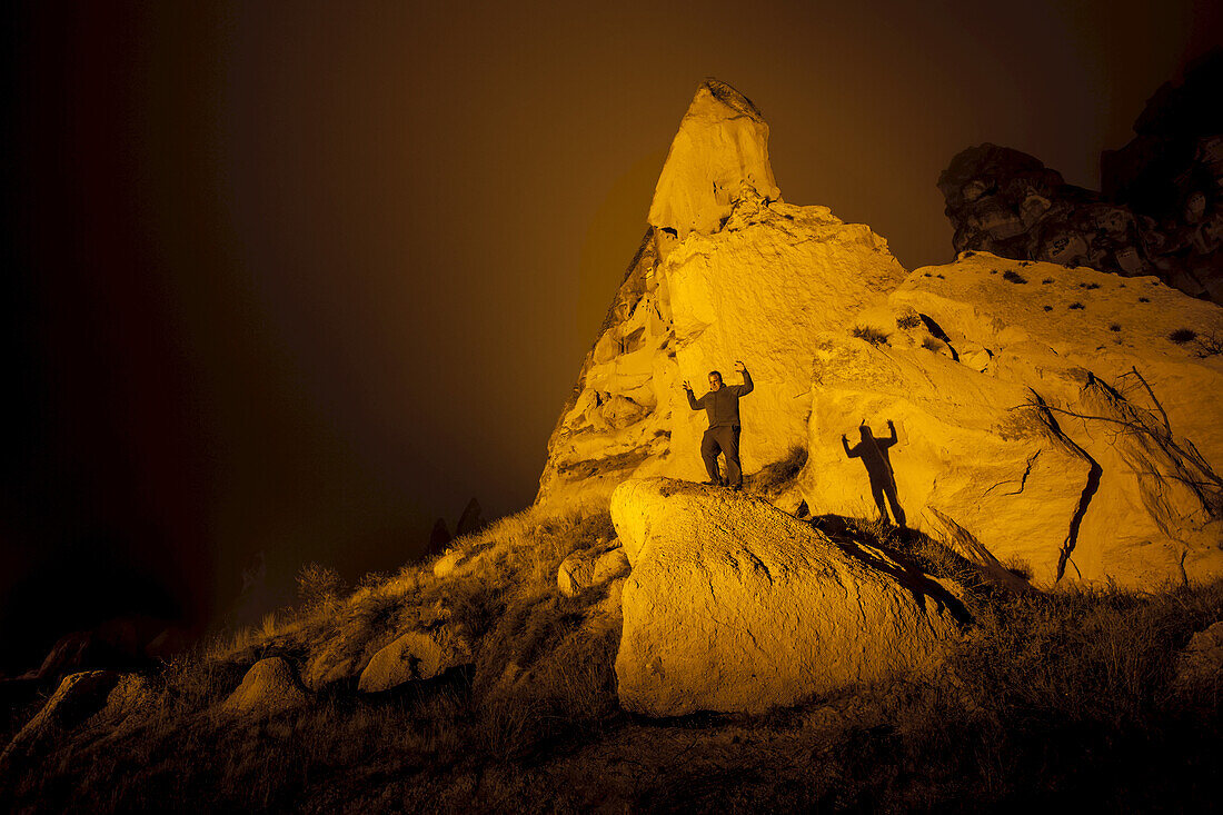 Ein Mann, der mit erhobenen Armen steht und einen Schatten auf Höhlenwohnungen in Felsformationen und Feenkamine wirft; Goreme, Kappadokien, Türkei