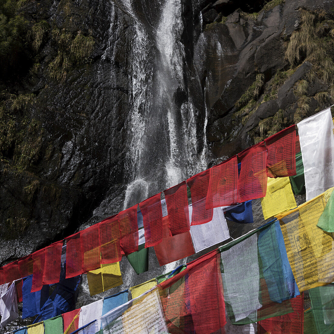 Bunte Gebetsfahnen und ein Wasserfall auf einer Klippe im Hintergrund; Paro, Bhutan