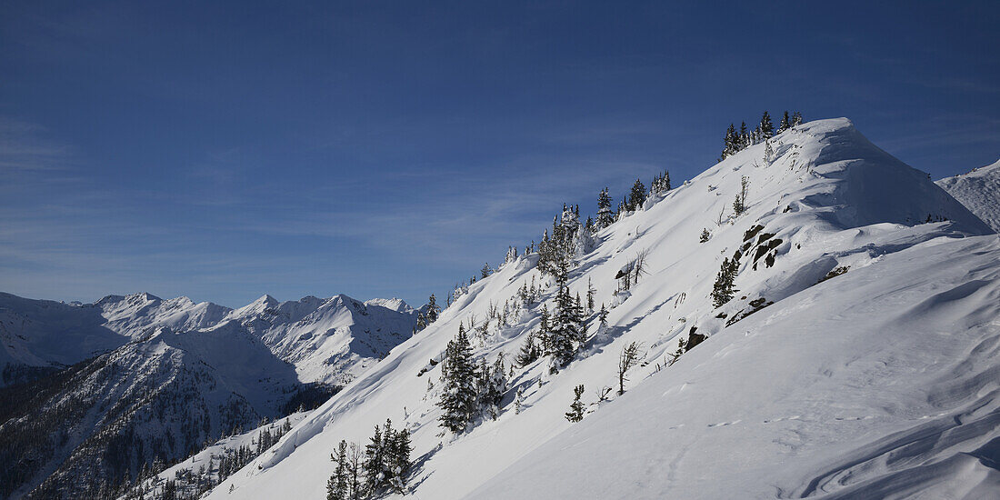 Verschneiter Berghang mit blauem Himmel und Blick auf eine Bergkette; Kicking Horse, British Columbia, Kanada