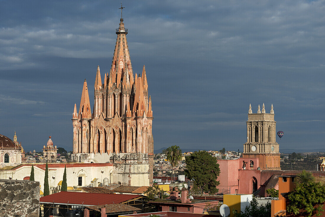 Kirche und Stadtbild unter bewölktem Himmel, mit einem Heißluftballon in der Ferne; San Miguel De Allende, Guanajuato, Mexiko