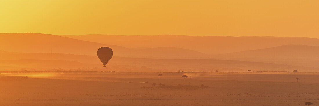 Ein Heißluftballon fliegt über die afrikanische Savanne im orangefarbenen Licht vor Sonnenaufgang, mit einer Staubspur von einem Lastwagen auf dem Boden; Narok, Kenia