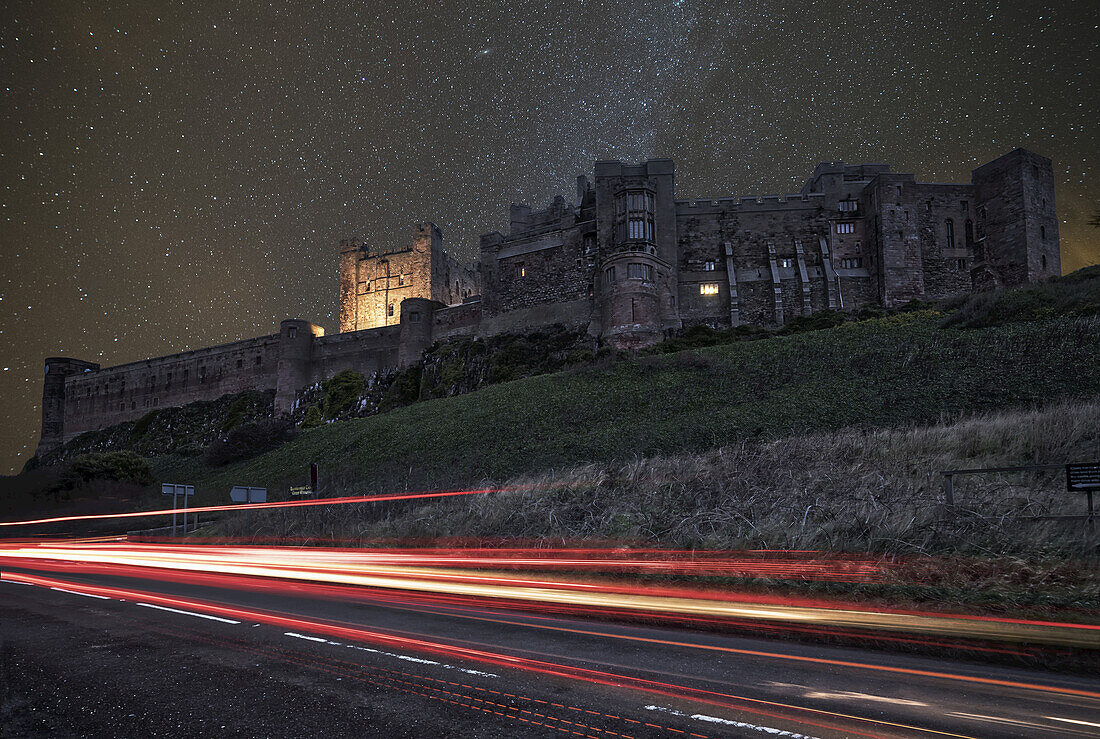 Lichtspuren auf einer Straße und ein sternenübersäter Himmel über Bamburgh Castle zur Nachtzeit; Bamburgh, Northumberland, England