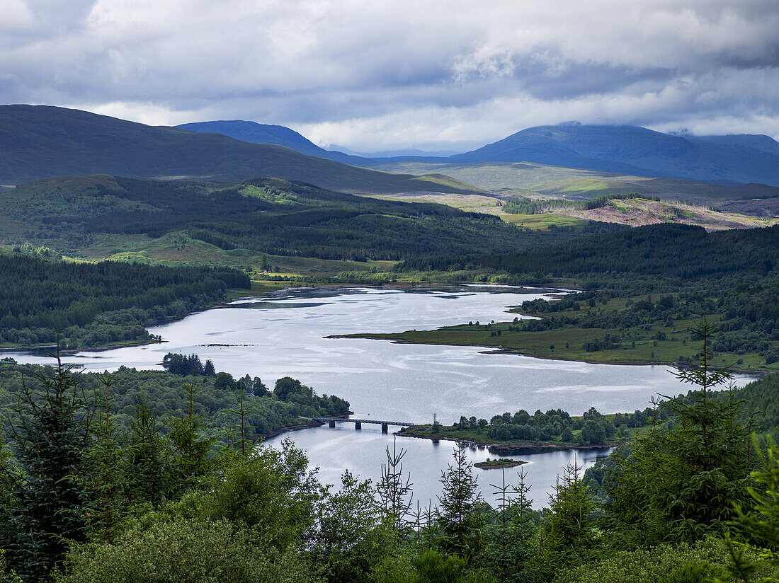 Flusslandschaft und Wälder über Bergen bei bewölktem Himmel; Schottland