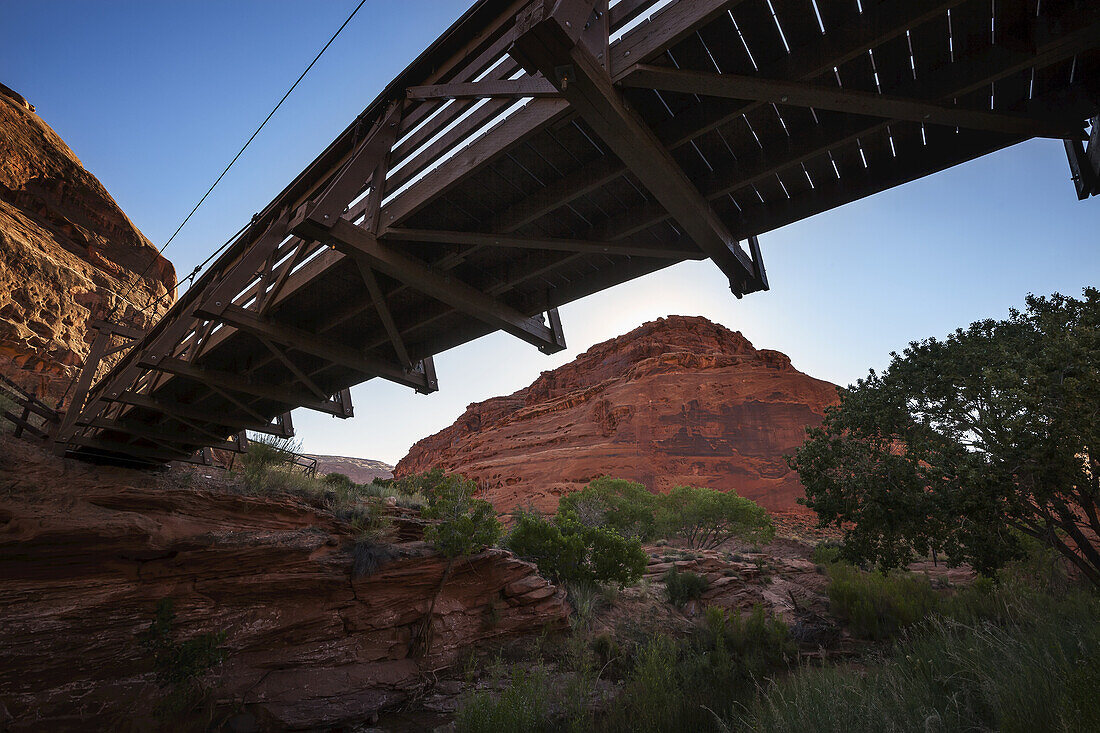 Eine Fußgängerbrücke, die einen Canyon-Bach überspannt, mit hohen Canyonwänden aus rotem Gestein im Hintergrund, von der Brücke aus gesehen; Utah, Vereinigte Staaten von Amerika