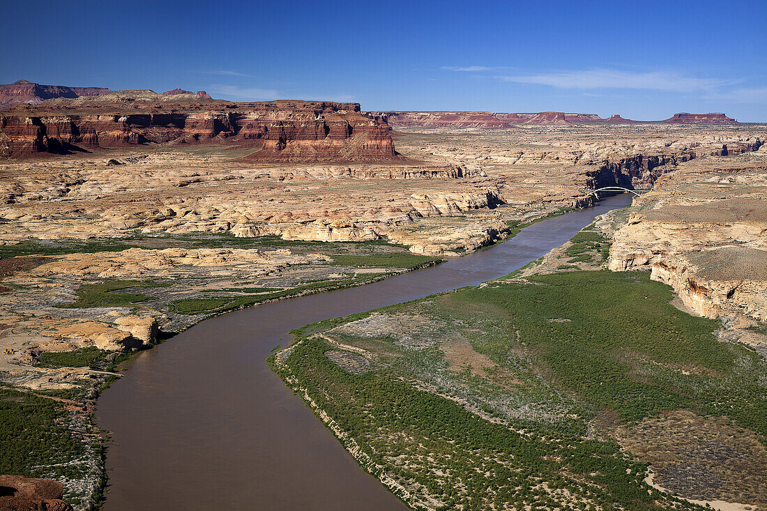 Luftaufnahme des Colorado River, der sich durch das Canyon Country von Süd-Utah schlängelt, mit einer Brücke, die den Fluss überspannt, im Hintergrund rote Canyonwände und blauer Himmel; Utah, Vereinigte Staaten von Amerika
