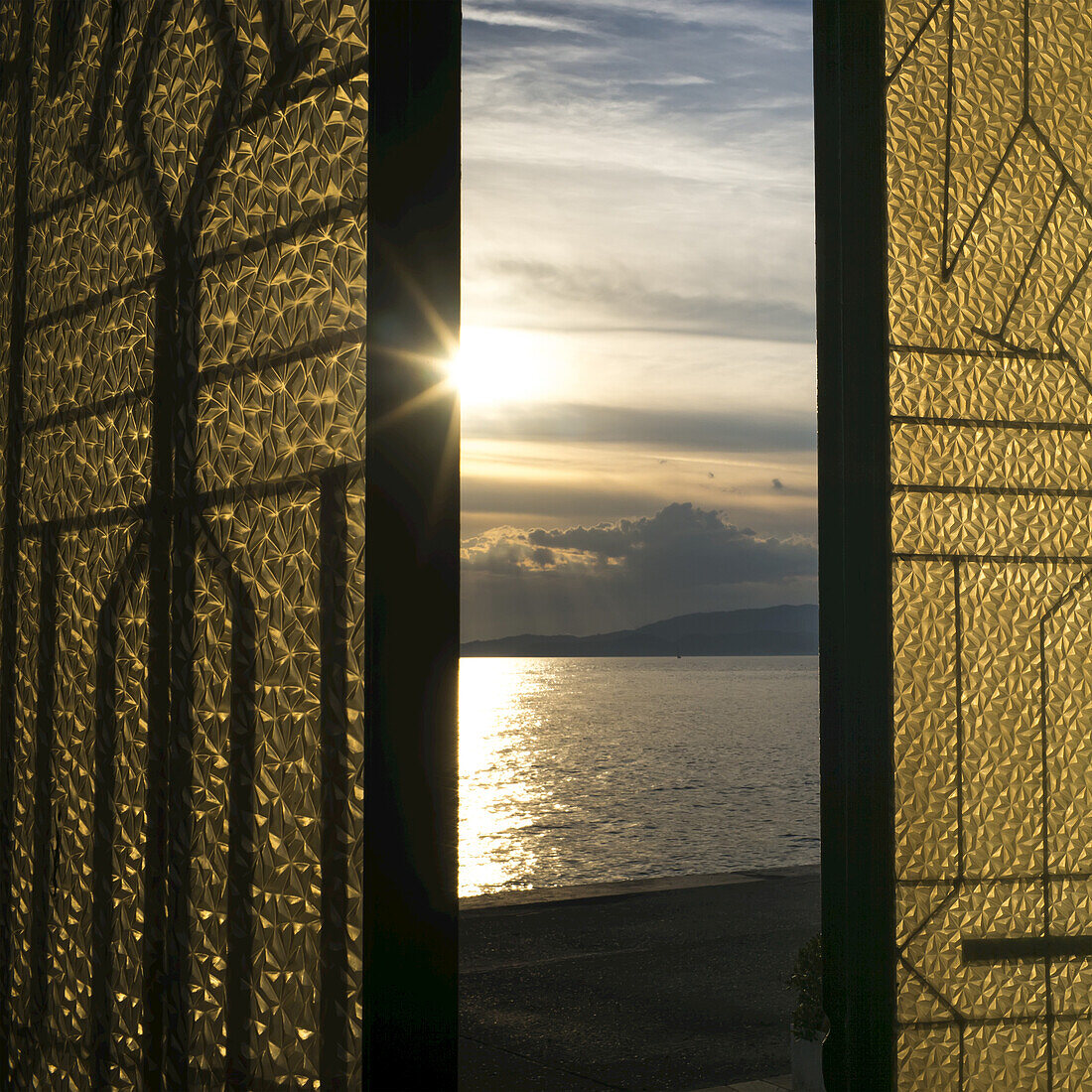 Sonnenlicht, das bei Sonnenuntergang leuchtet und sich im ruhigen Meerwasser spiegelt, betrachtet durch ein Fenster mit goldenen und schwarzen Fensterläden; Panormos, Thessalia Sterea Ellada, Griechenland