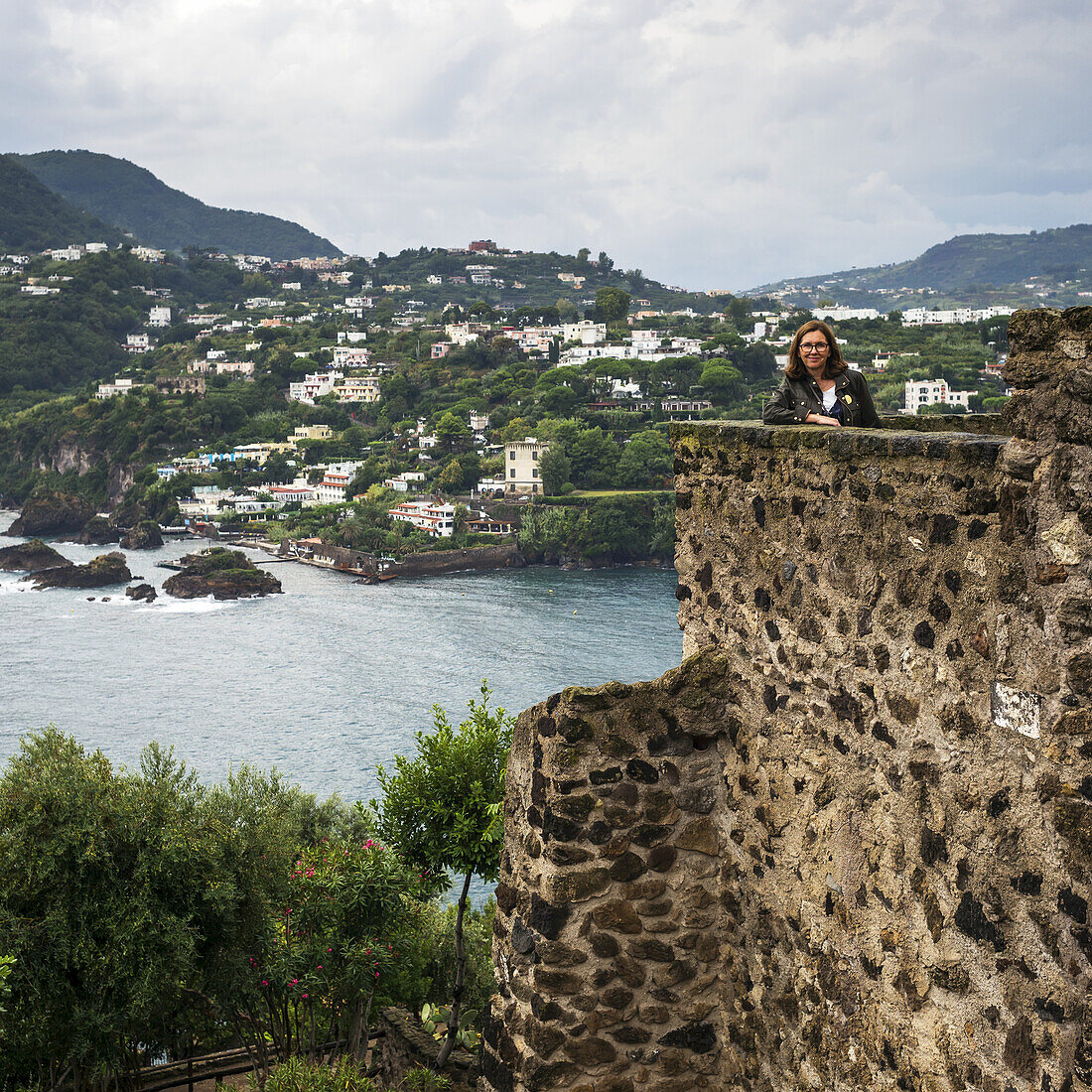 Eine Frau posiert vor einer alten Steinmauer des aragonischen Schlosses und blickt auf die Küste der Insel Ischia; Kampanien, Italien