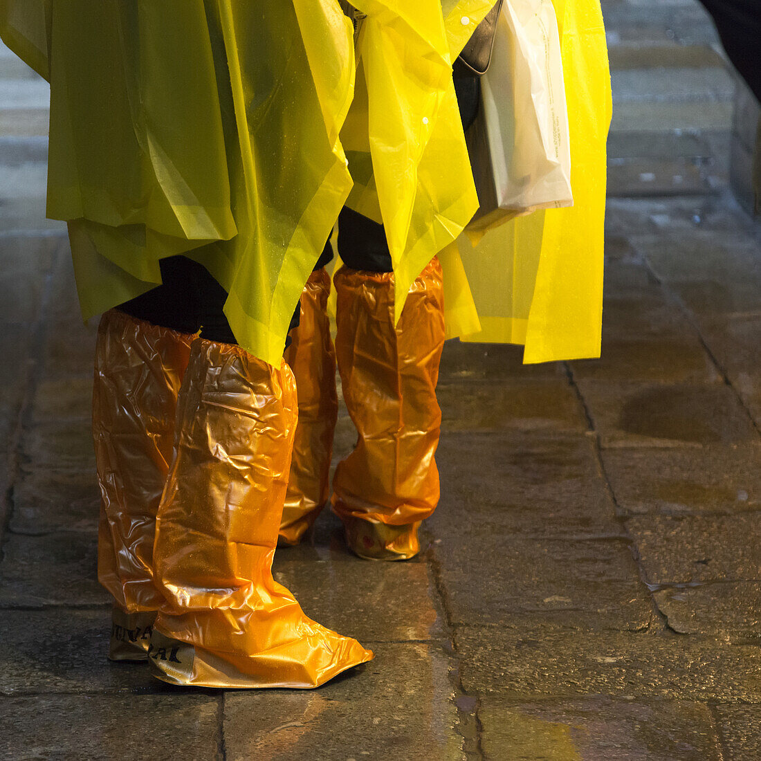 Stehende Menschen mit gelben Regenponchos und Schuhen, die mit orangefarbenen Plastikplanen bedeckt sind; Venedig, Italien