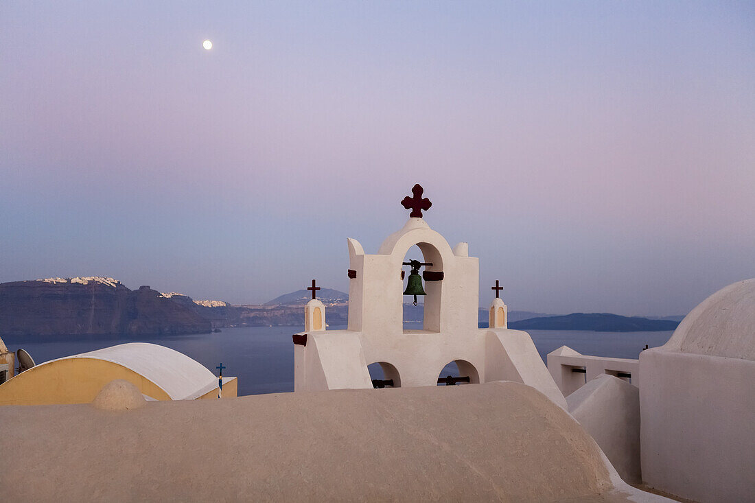 Orthodoxe Kirche mit Glocke und Mond hoch am Himmel in der Abenddämmerung; Oia, Santorin, Griechenland