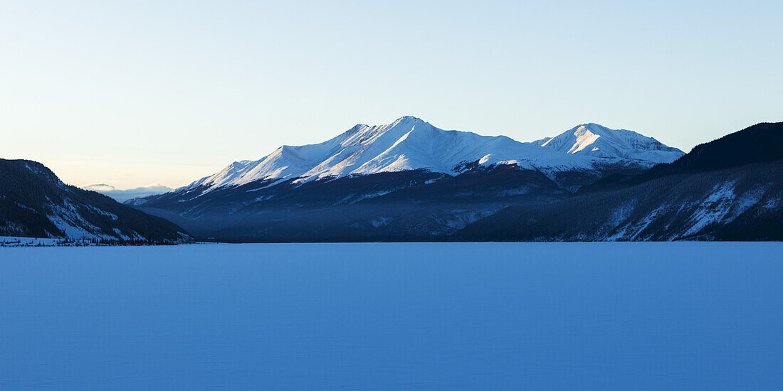 Frozen Snow Covered Lake With Snow Covered Mountains In The Background, Liard River Hot Springs Provincial Park; British Columbia, Canada