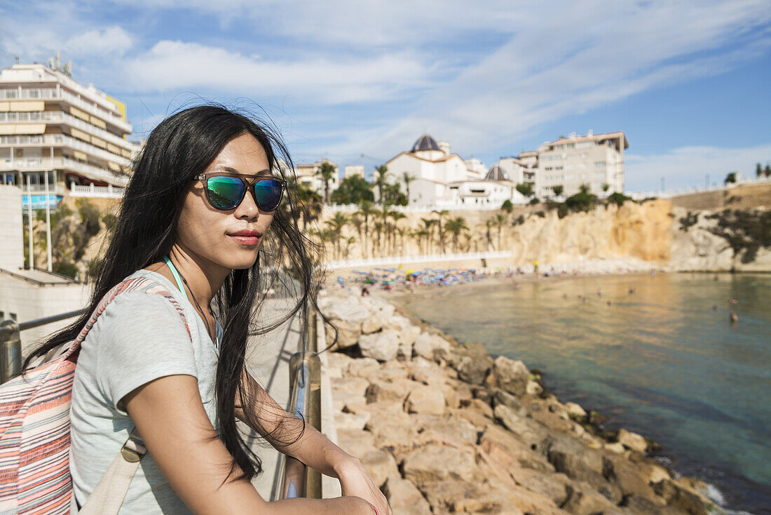 Eine chinesische junge Frau mit Sonnenbrille steht an einem Geländer und blickt auf das Wasser; Benidorm, Alicante, Spanien