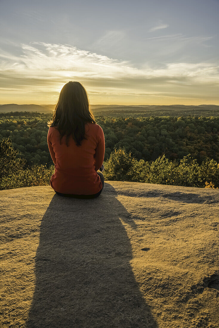 Frau sitzt auf einer Klippe mit Blick auf den Algonquin Park im Herbst bei Sonnenuntergang; Ontario, Kanada