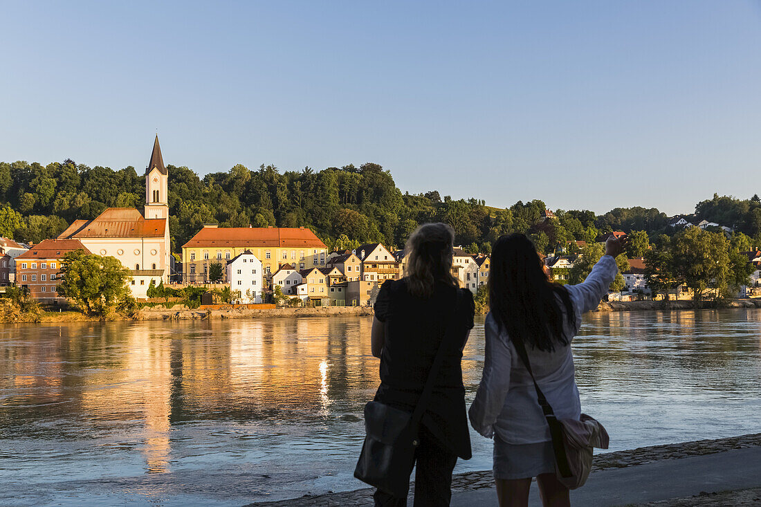 Water Reflects The Saint Gertrud Church On The River Inn, With Two Women Standing And Looking Away From The Camera In The Foreground Pointing Downriver; North Rhein-Westphali, Germany
