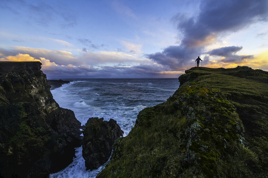 Person, die auf einer Klippenwand steht, während der Atlantik die Küste unter ihr stößt; Island
