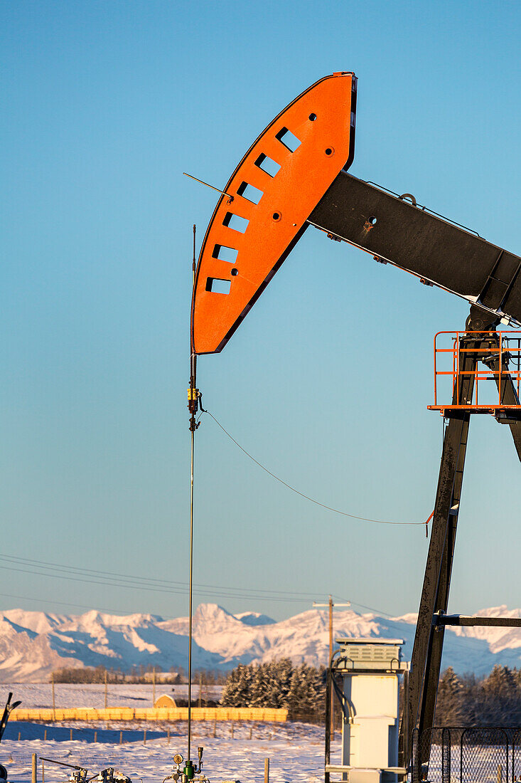 Close-up of pumpjack with the warm light at sunrise, snow-covered mountains and blue sky in the background, West of Airdrie; Alberta, Canada