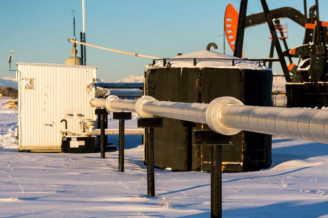 Close-up of metal pipeline, with a black metal tank, metal shed and pumpjack in the background in a snow-covered field, West of Airdrie; Alberta, Canada