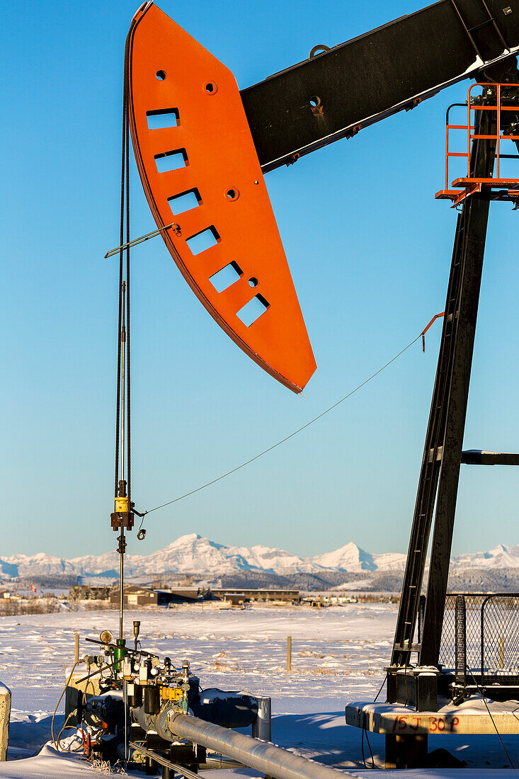 Close-up of pumpjack with snow-covered mountains and blue sky in the background, West of Airdrie; Alberta, Canada