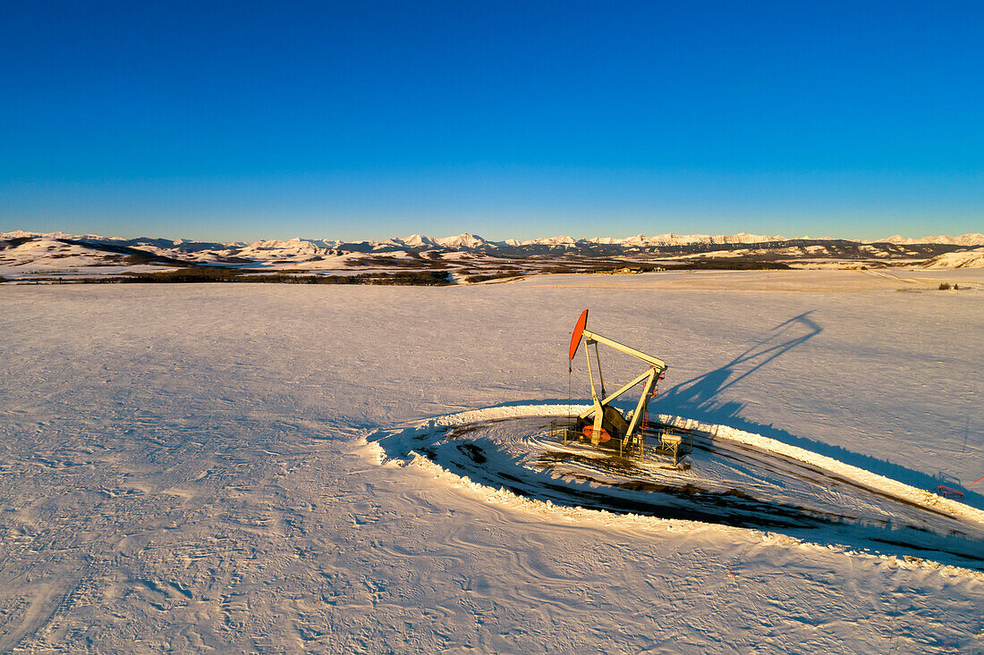 Aerial view of pumpjack in a snow-covered field with long shadows, warm light of sunrise, rolling hills, snow-covered mountains and blue sky in the background; Longview, Alberta, Canada