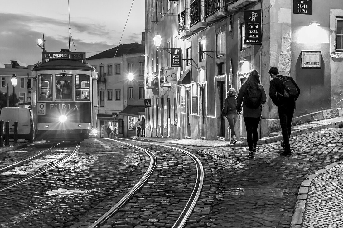 Straßenbahn und Fußgänger in der Abenddämmerung mit Lichtern, die das Straßenleben beleuchten; Lissabon, Region Lisboa, Portugal