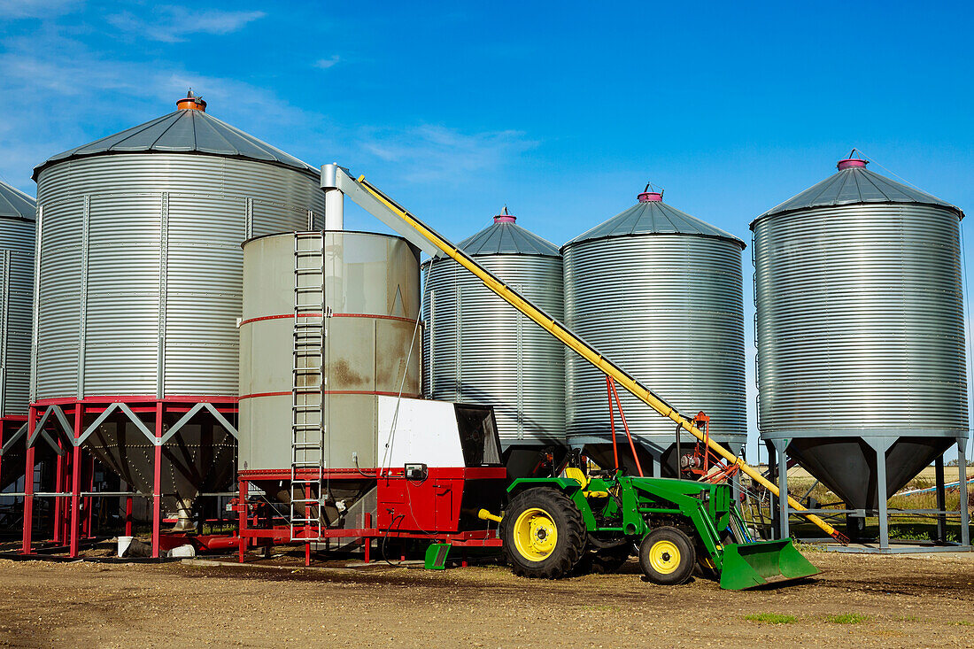 Silos in the background with a tractor and auger for loading a grain dryer in the foreground of a farm yard; Legal, Alberta, Canada