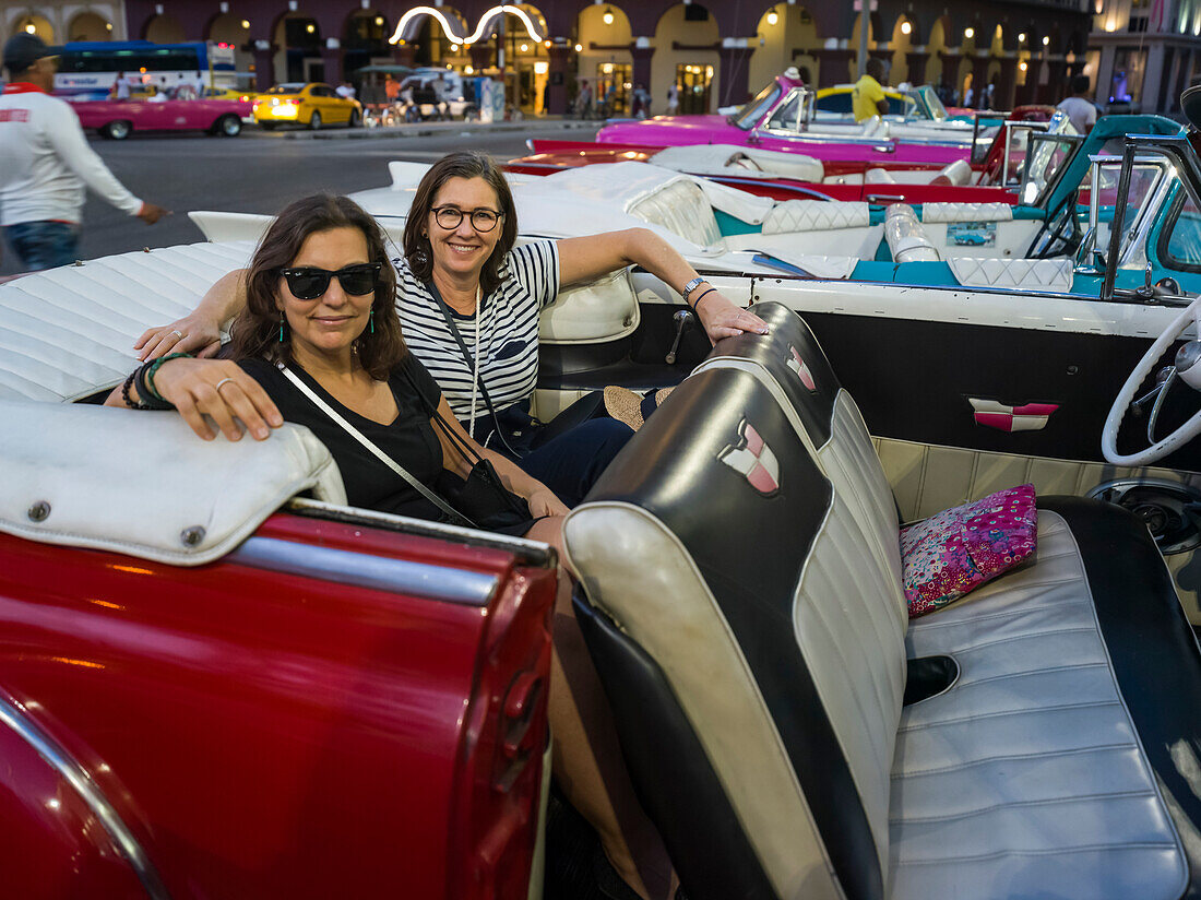 Two women sit in the backseat of a vintage car looking at the camera; Havana, Cuba