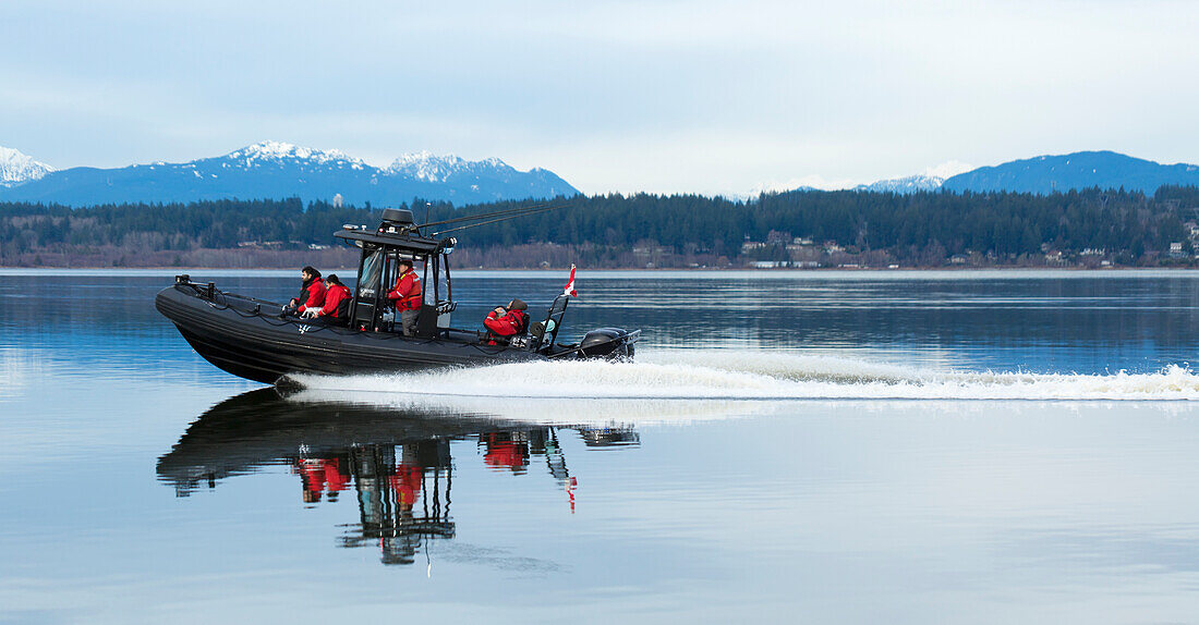 Motorboat with four people in red jackets riding along a coastline with the Coast Mountains in the background, Blackie Spit, Crescent Beach; Surrey, British Columbia, Canada