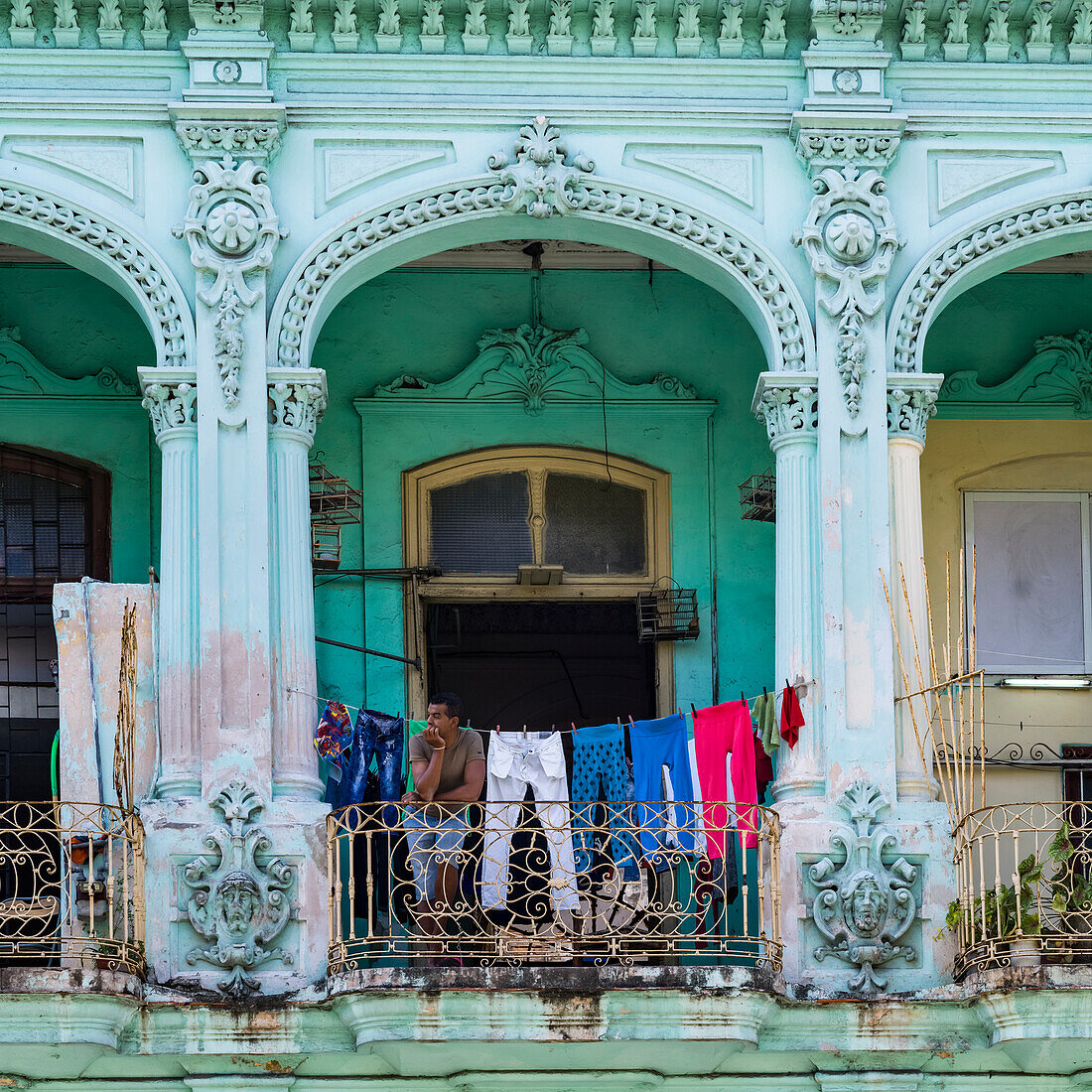 A building with ornate and decorative facade and a clothesline hanging across a balcony where a man sits looking out; Havana, Cuba