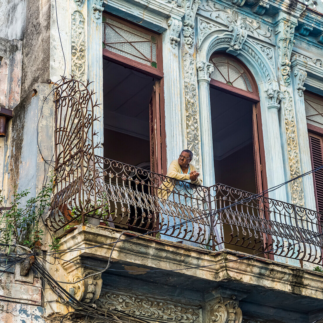 Frau lehnt am Geländer ihres Balkons und blickt auf die Straßen unter ihr; Havanna, Kuba