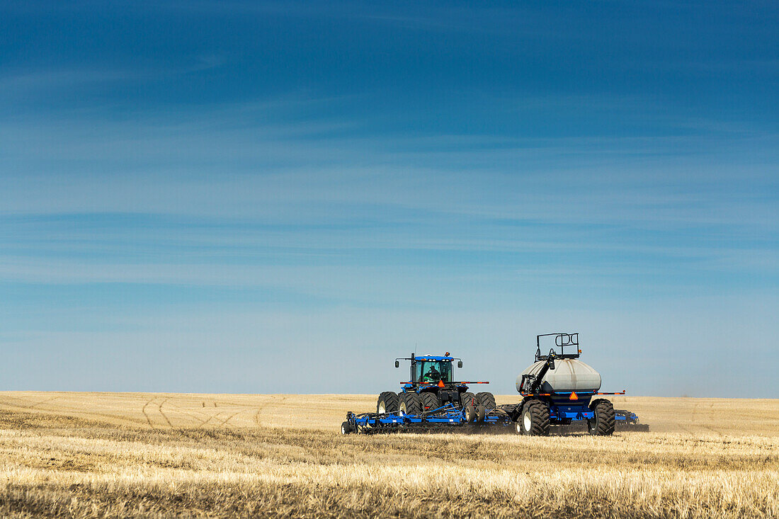 Tractor with air seeder in field with blue sky and hazy clouds, near Beiseker; Alberta, Canada