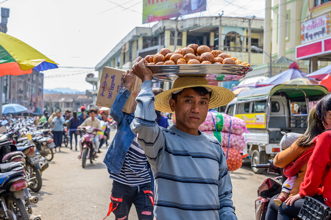 Junger Mann, der in einer belebten Straße Lebensmittel auf dem Kopf trägt; Lashio, Shan-Staat, Myanmar