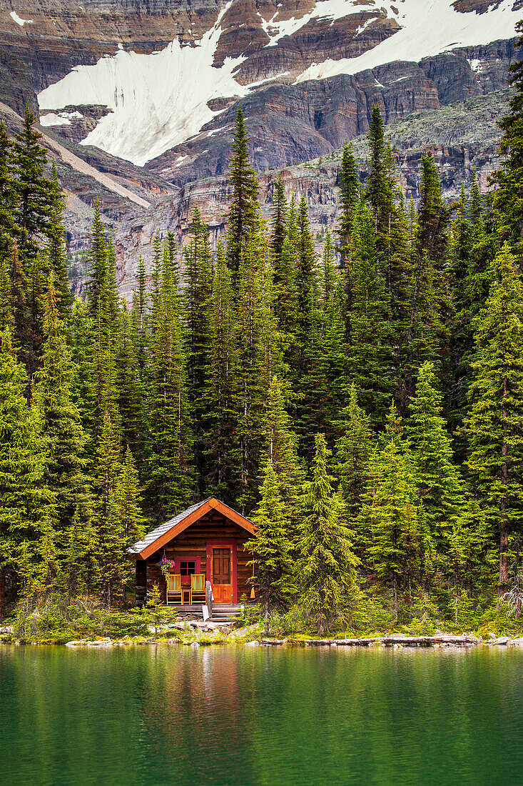 Holzblockhaus an einem Seeufer, das sich in der Sonne spiegelt, mit immergrünen Bäumen und Bergklippen im Hintergrund; Field, British Columbia, Kanada