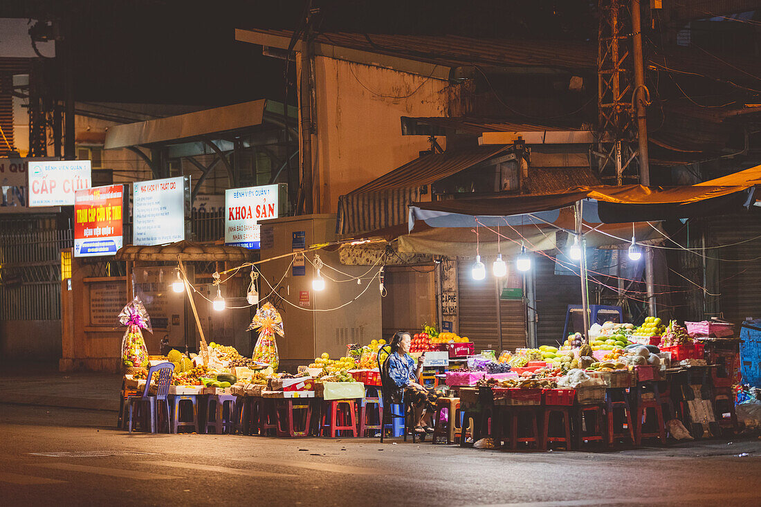 A senior woman vendor sits using her cell phone at a night market; Ho Chi Minh City, Vietnam