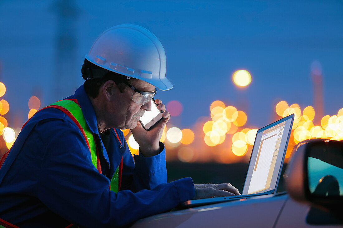 Man working on a laptop and smart phone with an oil refinery in the background; Alberta, Canada