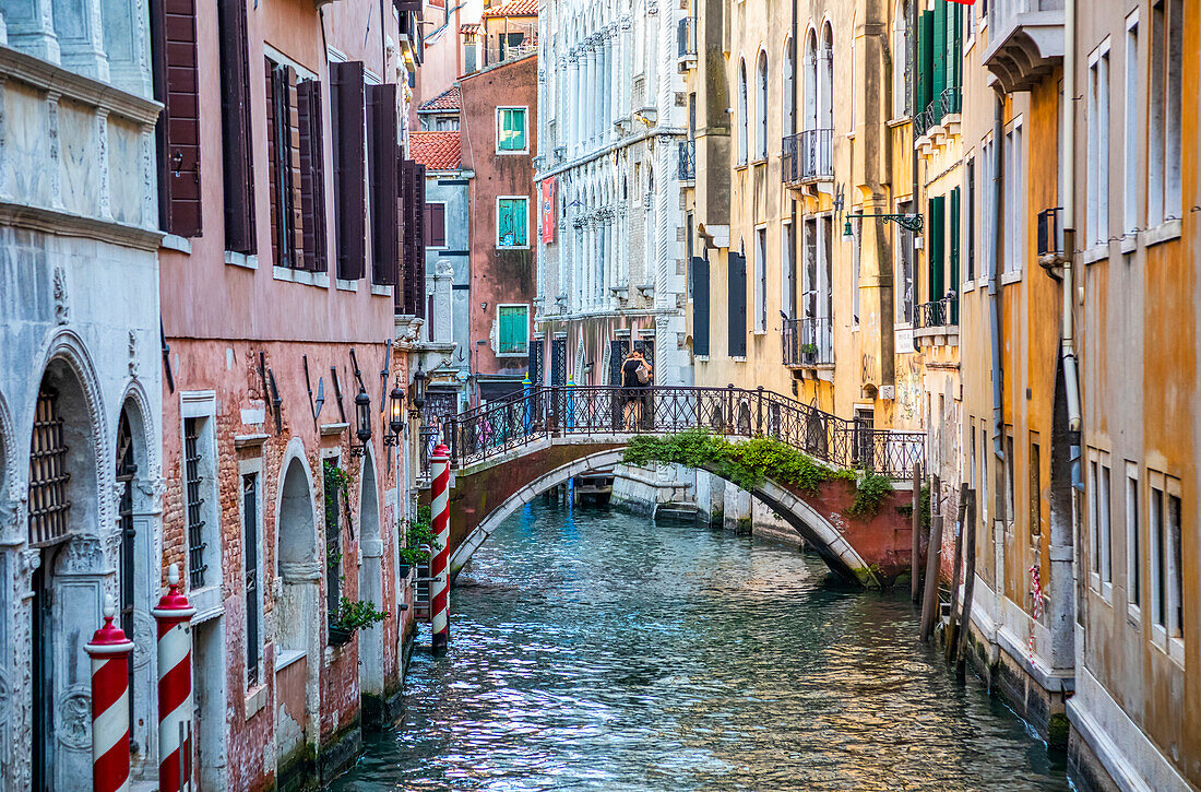 Ein Paar umarmt sich auf einer bogenförmigen Fußgängerbrücke über einen Kanal, mit bunten Gebäuden auf beiden Seiten; Venedig, Italien