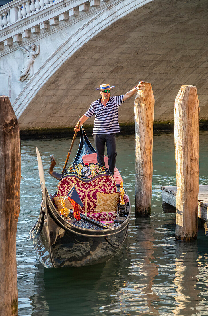 Gondolier stands on the back of a gondola at the waterfront waiting for tourists; Venice, Italy