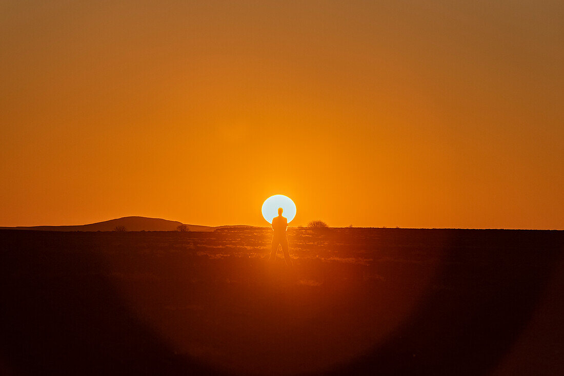 Ein Mann steht im Gegenlicht der hellen, glühenden Sonne, die bei Sonnenuntergang hinter dem Horizont versinkt, in Aluvlei, Namib-Naukluft-Nationalpark; Namibia