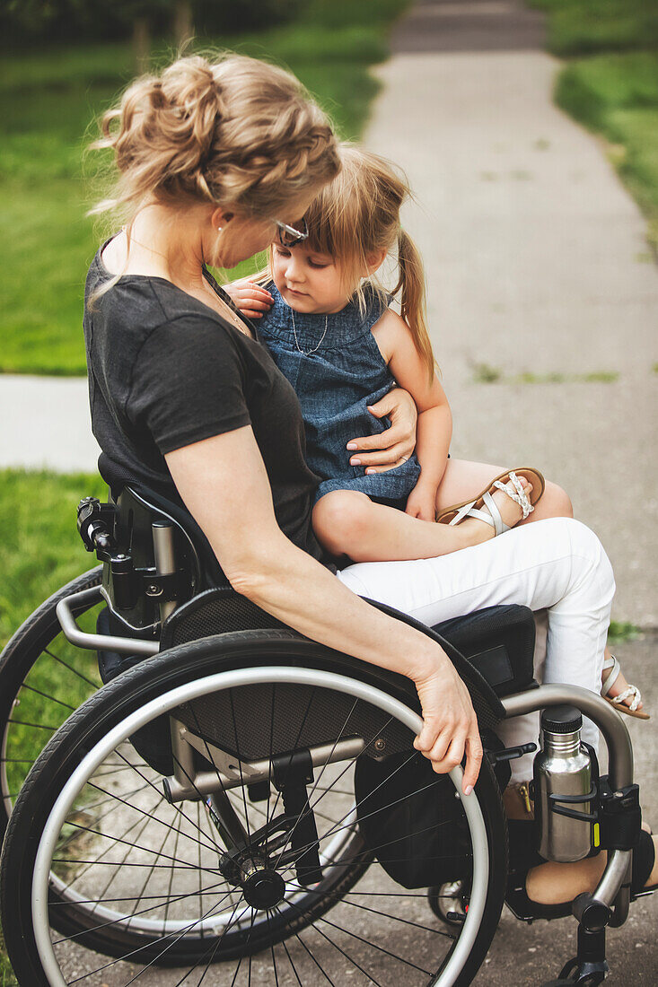 Eine querschnittsgelähmte Mutter, die ihr kleines Mädchen auf dem Schoß hält, während sie es an einem warmen Sommernachmittag in ihrem Rollstuhl die Straße hinunterfährt: Edmonton, Alberta, Kanada.