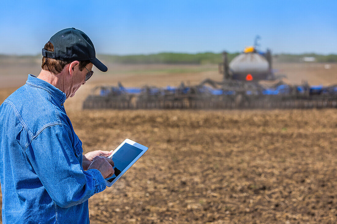 Landwirt mit Tablet, während er auf einem landwirtschaftlichen Feld steht und ein Traktor mit Geräten das Feld besät; Alberta, Kanada