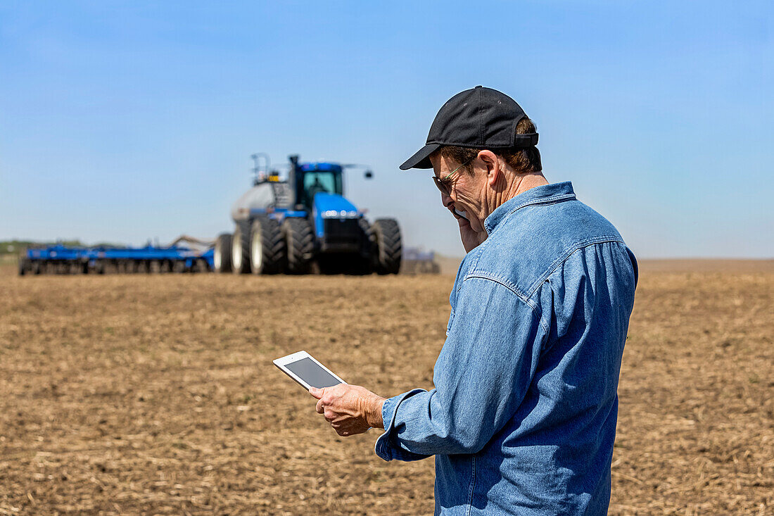 Farmer using a smart phone and tablet while standing on a farm field and watching the tractor and equipment seeding the field; Alberta, Canada