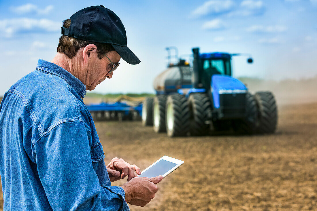Landwirt, der ein Tablet benutzt, während er auf einem landwirtschaftlichen Feld steht und einen Traktor und Geräte beobachtet, die das Feld säen; Alberta, Kanada