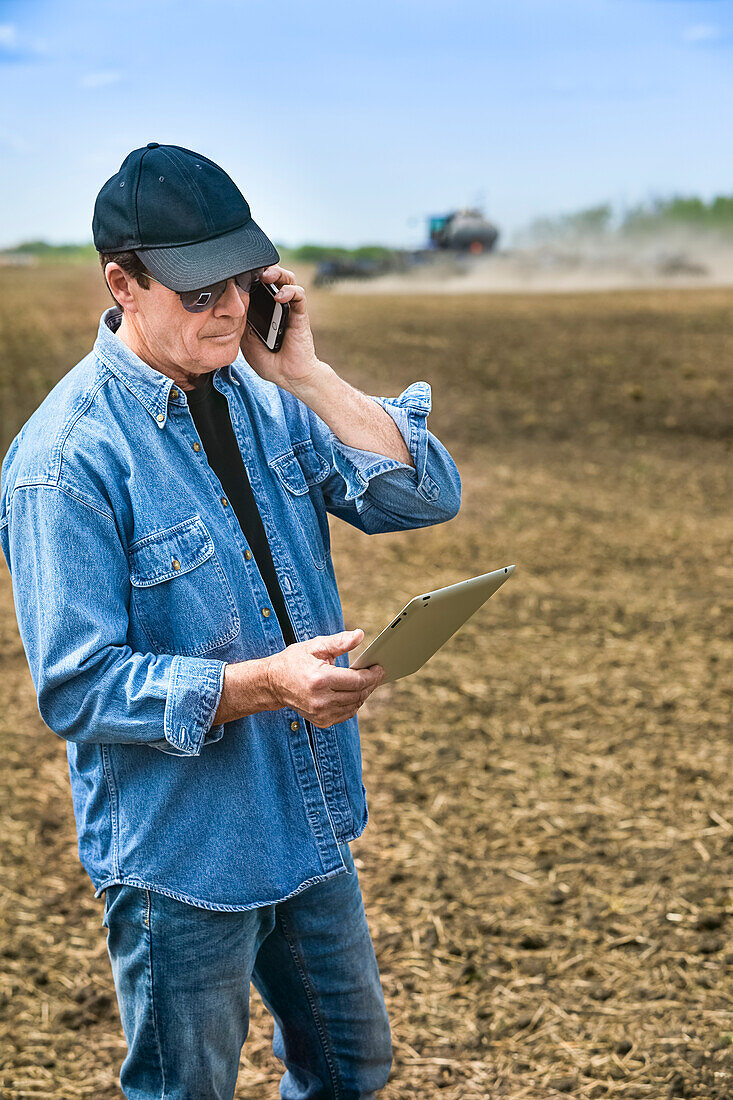 Landwirt, der ein Smartphone und ein Tablet benutzt, während er auf einem landwirtschaftlichen Feld steht und den Traktor und die Maschinen beim Säen des Feldes beobachtet; Alberta, Kanada