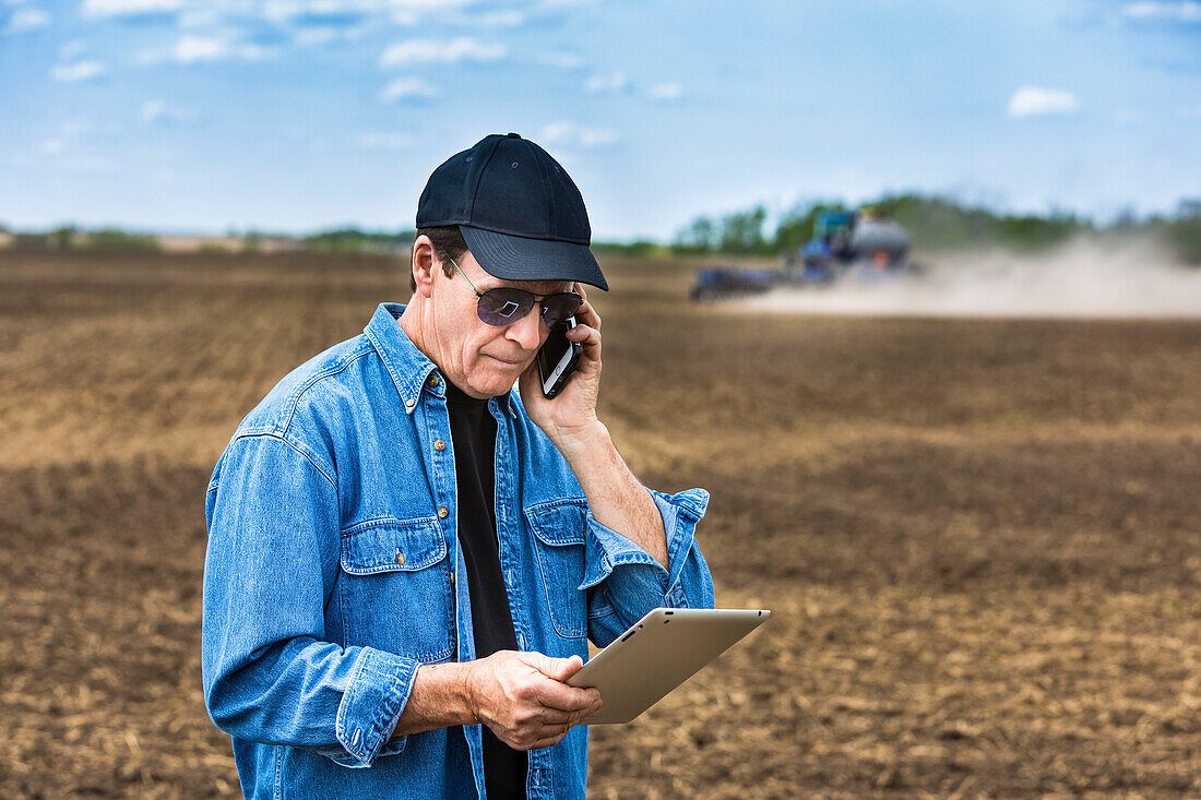 Ein Landwirt benutzt ein Smartphone und ein Tablet, während er auf einem landwirtschaftlichen Feld steht und den Traktor und die Geräte bei der Aussaat beobachtet; Alberta, Kanada