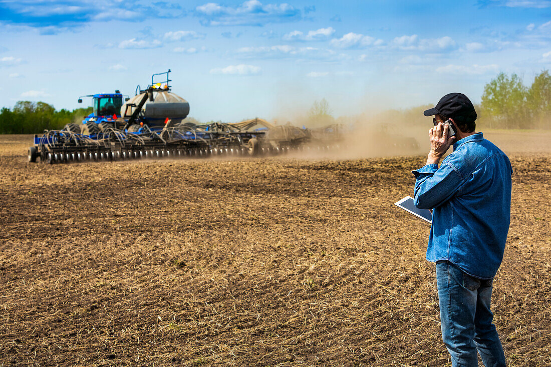 Farmer using a smart phone and tablet while standing on a farm field and watching the tractor and equipment seeding the field; Alberta, Canada