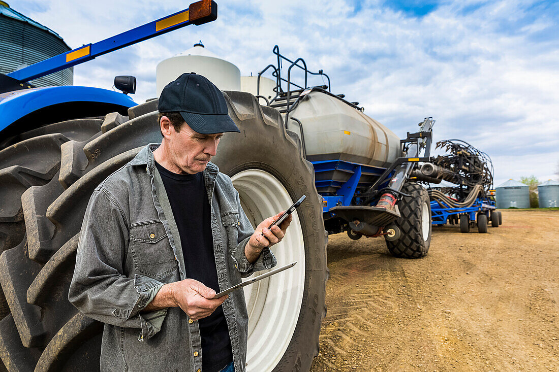 Farmer using a smart phone and tablet while standing on a farm beside equipment; Alberta, Canada